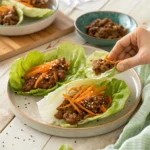 Ground Beef Lettuce Wraps That Actually Taste Amazing 9 A hand picking up one of several beef lettuce wraps topped with carrots and sesame seeds on a ceramic plate